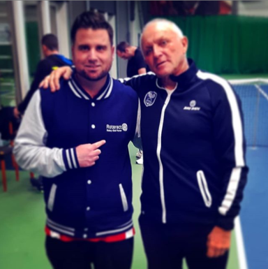Zwei Männer in Sportjacken posieren freundlich auf einem Tennisplatz.